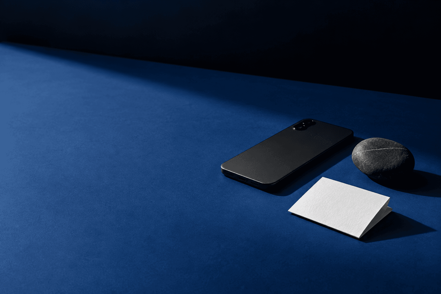 A black smartphone face down beside a white note and stone on a blue surface